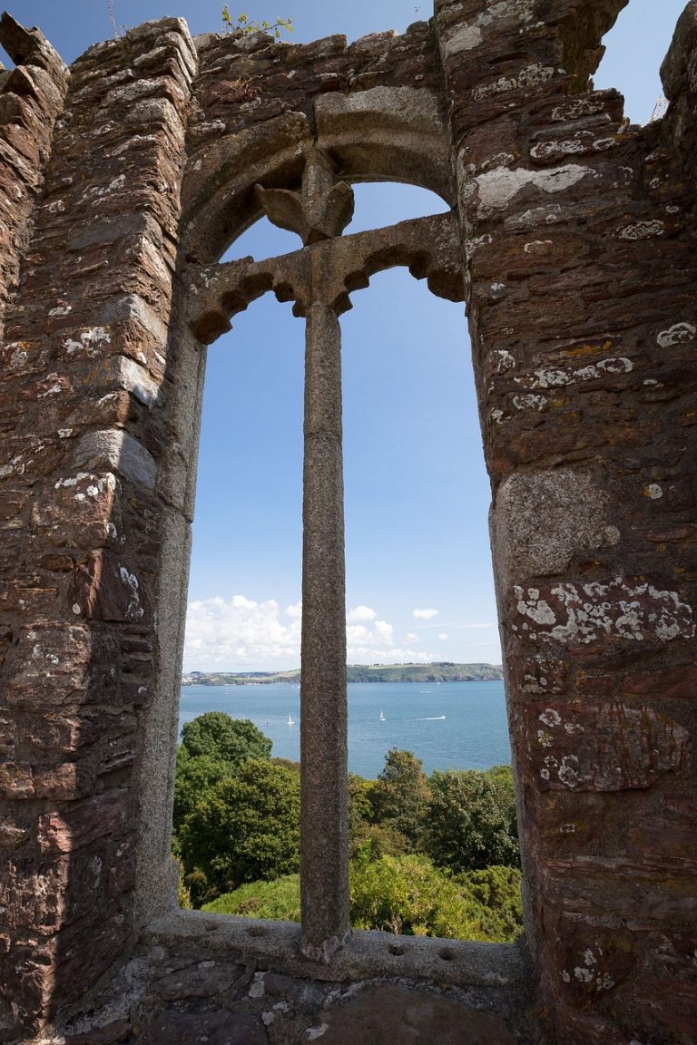 View through a stone arched window ruin out to a coastal landscape with trees, water, and sailboats under a blue sky